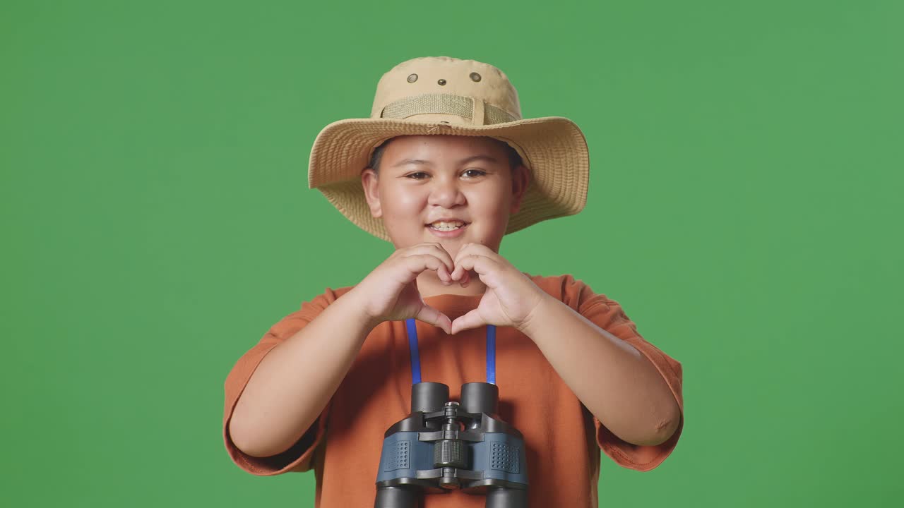 niño turista asiático con un sombrero y binoculares haciendo un gesto en forma de corazón mientras está de pie en el fondo de la pantalla verde. niño investigador examina algo, concepto de aventura de turismo de viaje, de cerca