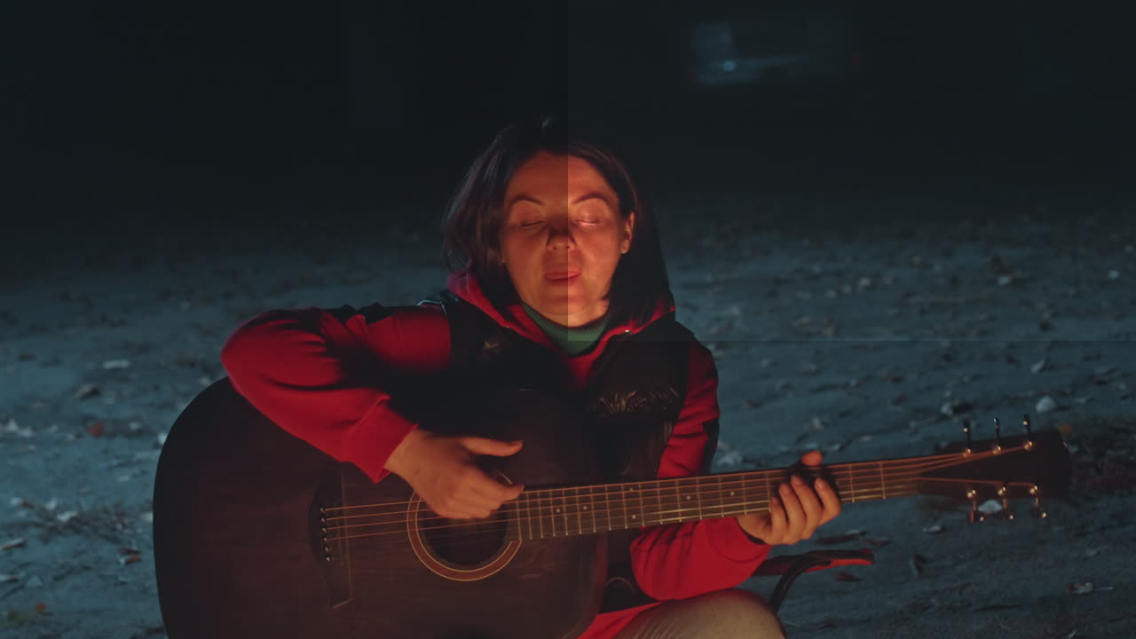 Donna bianca che suona la chitarra in campeggio di notte, che canta accanto al fuoco da campo, che indossa una giacca rossa, viso espressivo, strimpellando accordi costanti, melodia folk rilassante, esibizione intima all'aperto.