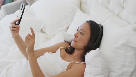 Happy-asian-woman-lying-in-bed,-wearing-headphones-and-using-smartphone,-in-slow-motion