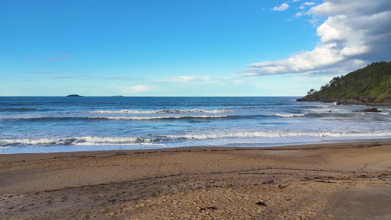 Gentle waves roll onto the sandy shore under a clear blue sky at Charlesworth Bay Beach, Coffs Harbour