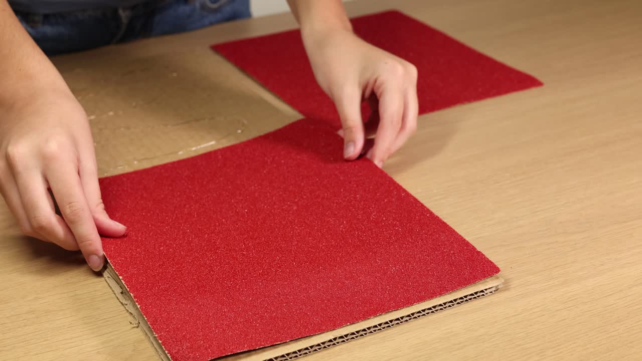 Hands placing and pressing red sandpaper on a cardboard surface under bright lighting, focusing on texture and material interaction