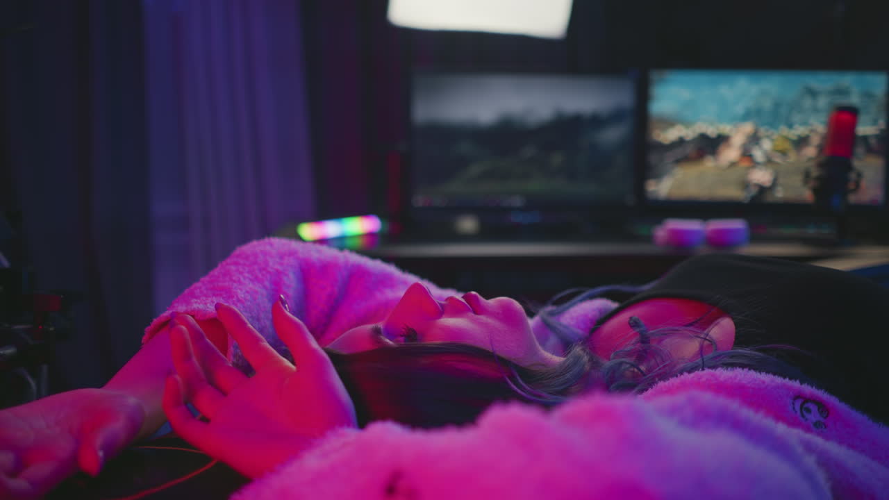 Side view of young woman sleeping in gaming chair under pink and purple neon light, eyes closed and arms relaxed, with computer monitors and studio setup in background
