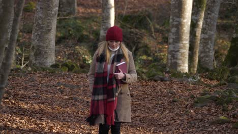 mujer joven con sombrero rojo y bufanda sostiene un libro caminando en el bosque de otoño