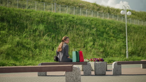 lady approaches bench, places colorful shopping bags on it, sits down, and relaxes her left hand while moving her left leg forward, she gazes into the distance with a green hill in the background