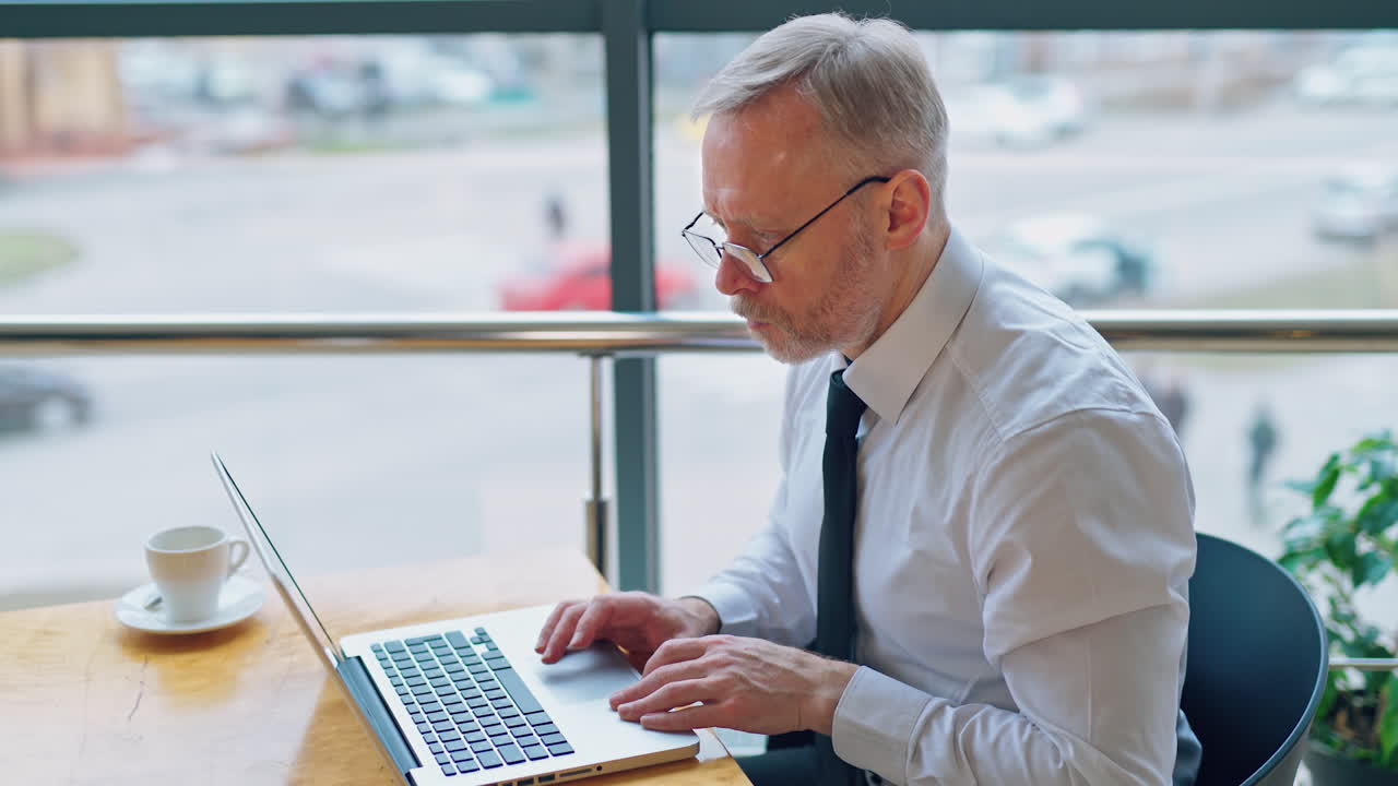 Freelance job. Mature man working on a laptop near the window. Serious businessman typing on a computer indoors on city background.