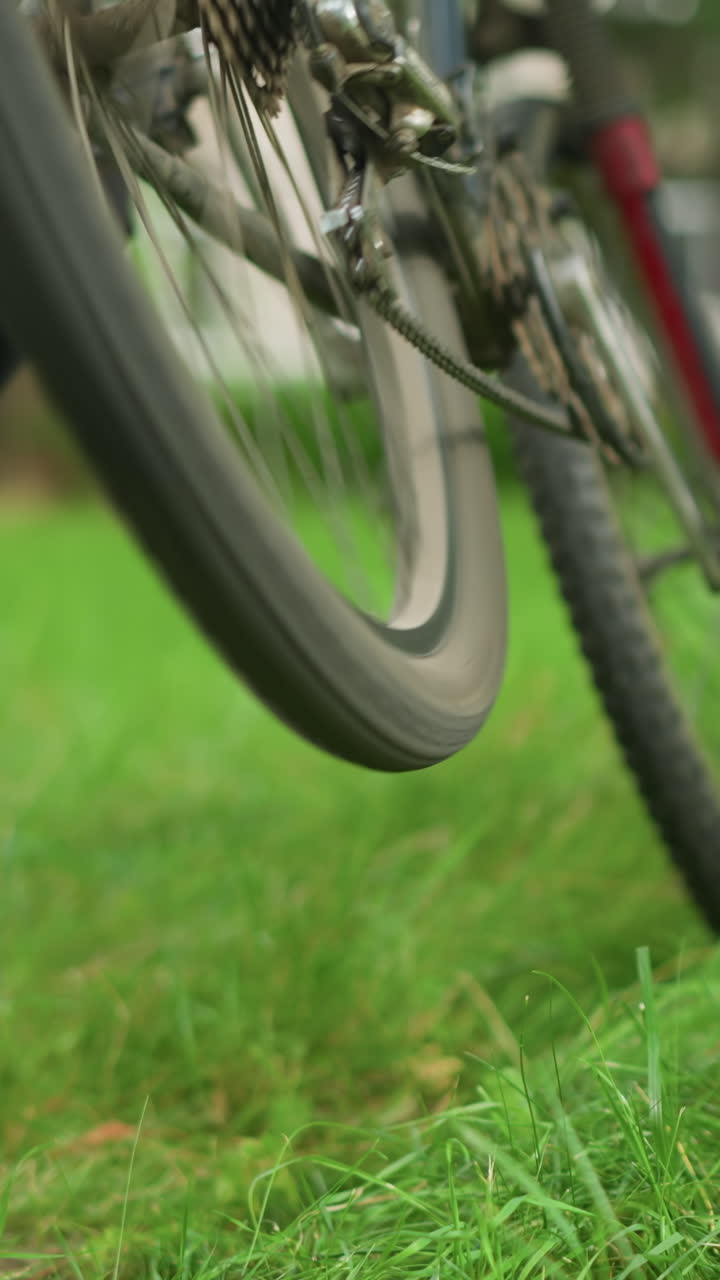 vista de la pierna de alguien con pantalones negros y zapatillas de deporte caminando hacia una bicicleta estacionada en un campo cubierto de hierba, ajusta el pedal, levanta los neumáticos traseros y pedala, haciendo que los neumáticos giren brevemente