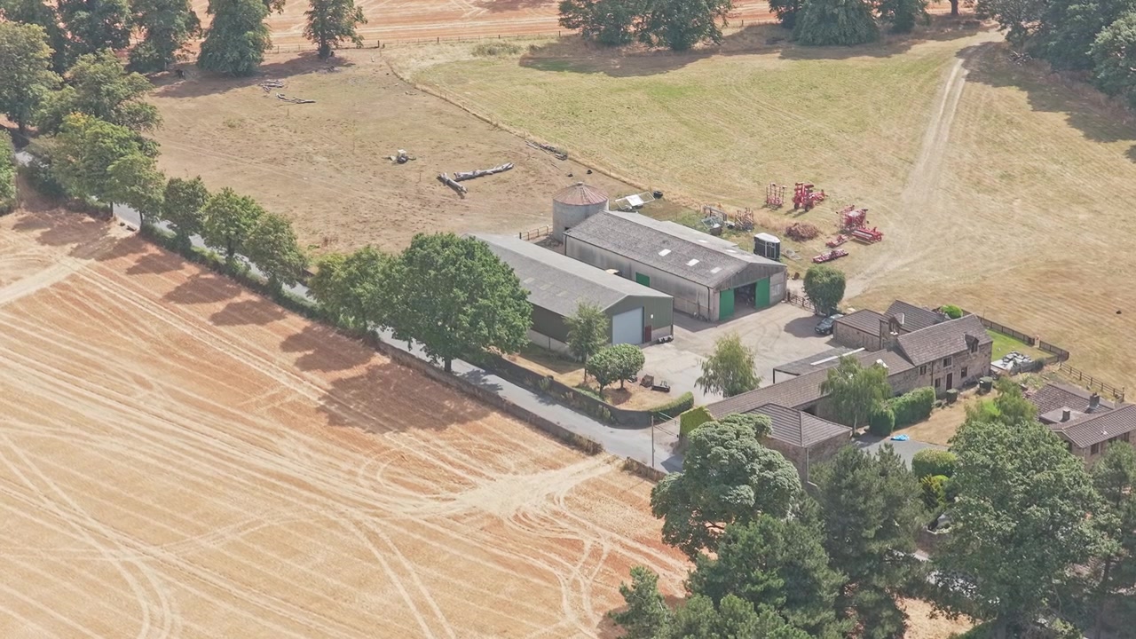 Propriedade agrícola situada ao lado de campos cultiváveis, sebes e bosques perto de Newmillerdam, West Yorkshire, com edifícios agrícolas, maquinário e trilhas de campo, sobrevoo de drone para frente