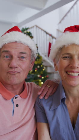 una foto vertical de una pareja caucásica mayor haciendo una llamada de video, con sombreros de papá noel.
