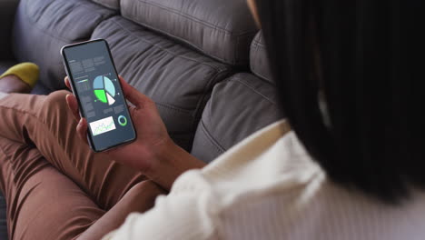 mujer biracial sentada en el sofá usando un teléfono inteligente con estadísticas en la pantalla