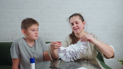 mother carefully unfolding handcrafted paper ornament while son watches closely with serious expression sitting together indoors at glass table against white brick wall decorated with simple cushions