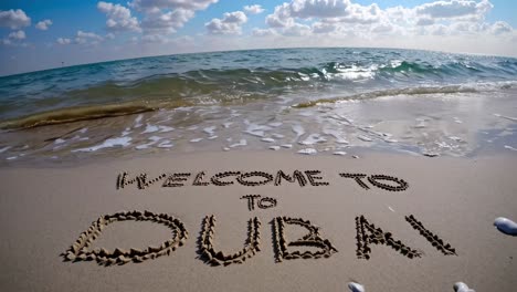 turquoise sea water gently caressing the shore, where someone has written welcome to dubai in the sand, all beneath a stunning, cloudy sky