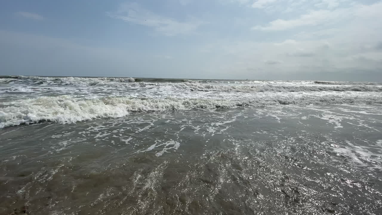 Panoramic view of small waves splashing at the shore of an empty beach. In the back of the scene is the ocean's horizon. Sunny day with some clouds.