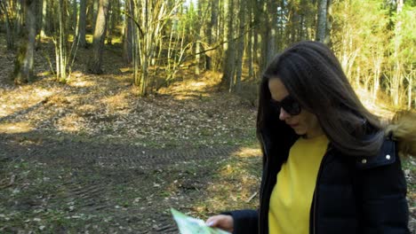 joven mujer caucásica con cabello castaño y gafas de sol oscuras caminando en la madera, mapa en la mano, hermoso día soleado de primavera, seguimiento de fosa tomada desde el suelo