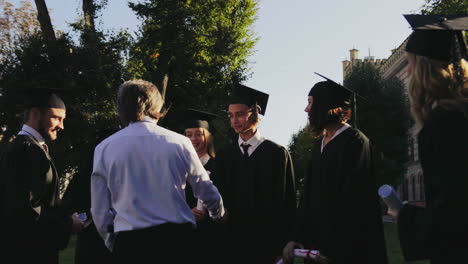 Gray-haired-middle-aged-proffessor-shaking-hands-and-hugging-multi-ethnical-graduates-after-the-graduation-ceremony