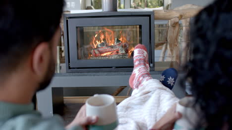 Glückliches-Biracial-Paar-Auf-Dem-Sofa-In-Warmen-Socken-Unter-Der-Decke-Vor-Dem-Kamin-Zu-Hause,-Zeitlupe