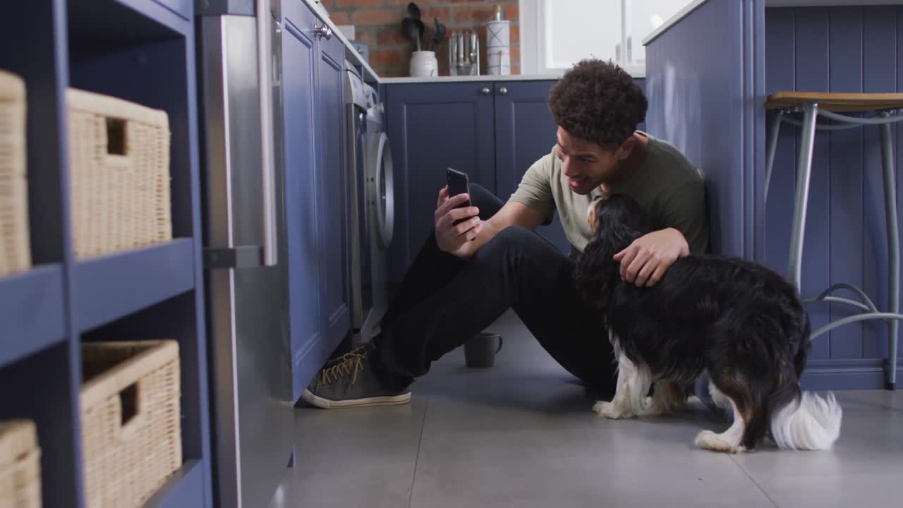 Happy biracial man sitting on floor in kitchen with his pet dog, making video call using smartphone