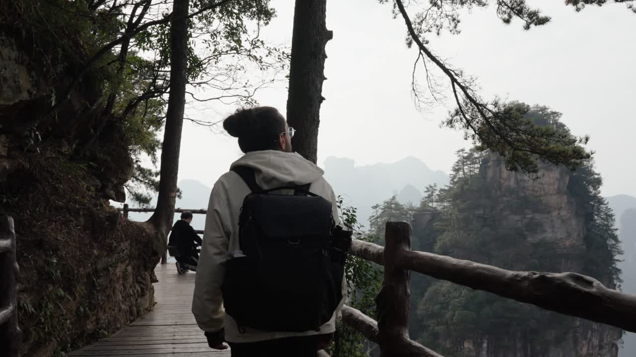 A man with a backpack walks a wooden trail carved along a cliff in Zhangjiajie, surrounded by misty forest and tall rock pillars.