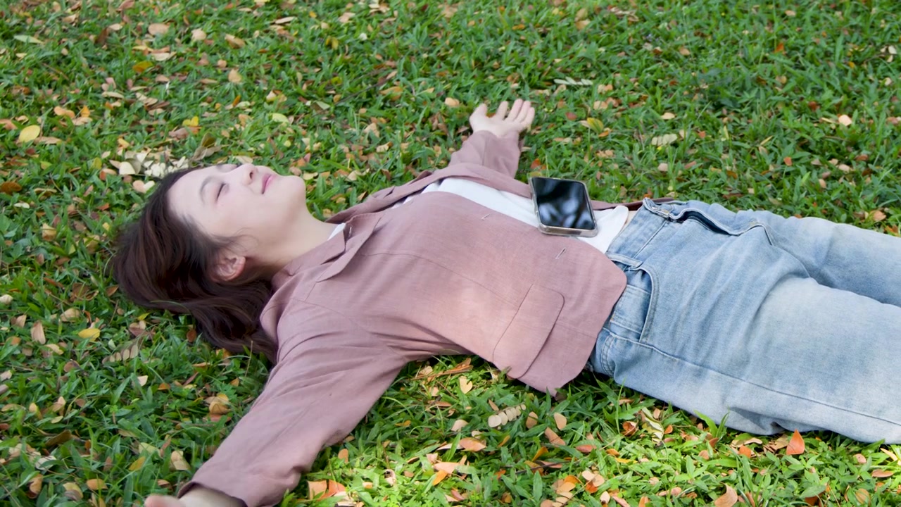 Young woman in pink blazer rests on lawn, eyes closed, under bright natural sunlight