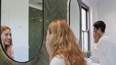 Happy-caucasian-lesbian-couple-looking-in-mirror,-touching-face-and-smiling-in-bathroom