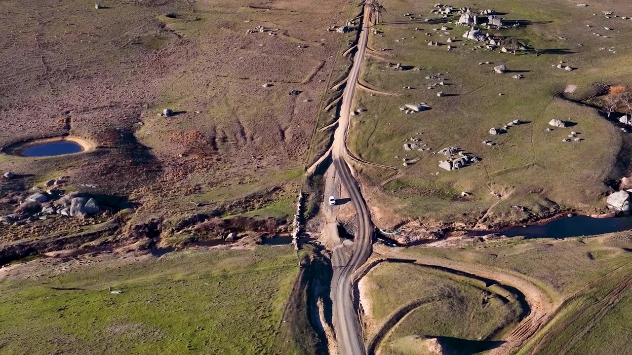 Ripresa aerea con drone dall'alto di un veicolo bianco che viaggia lungo una strada sterrata tortuosa nell'entroterra.
