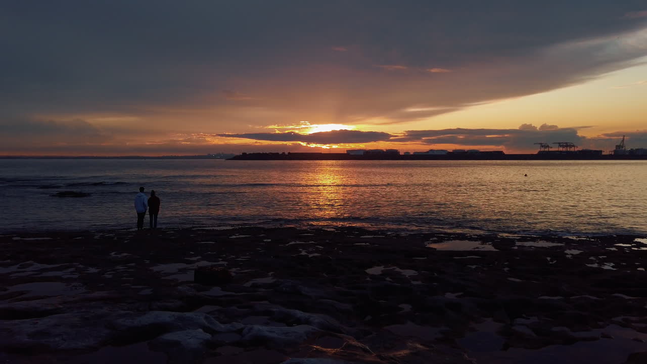 Couple stands romantically on rocky shore looking at colorful sunset on horizon