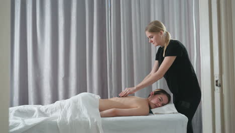 massage therapist in black attire using stone to gently massage client back on white table in serene wellness room with soft natural light and gray curtain backdrop, creating a calm environment