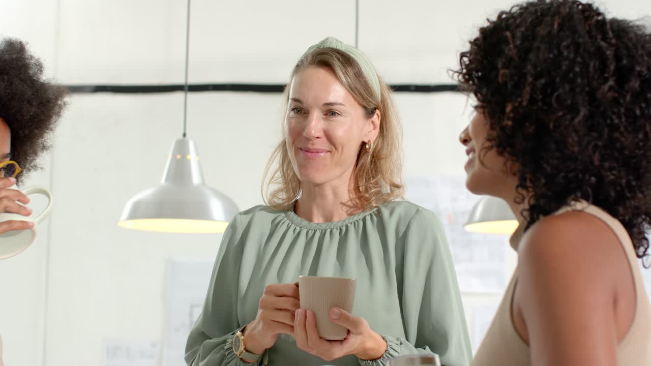 Diverse women coworkers standing at office break holding mugs, left raising mug, center speaking