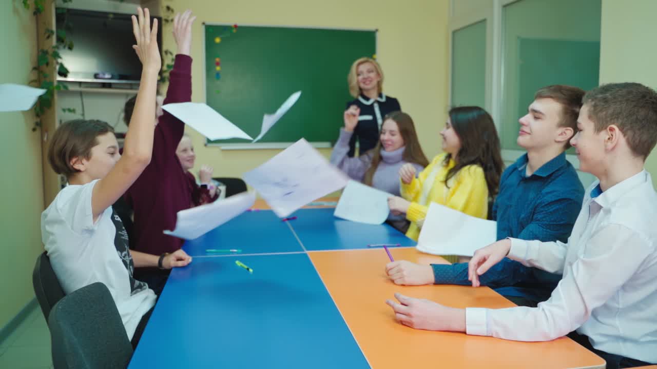 Teen students at school. Happy children sitting in the classroom and throw papers up. Students enjoys the time together. Papers flying in the class. Slow motion.
