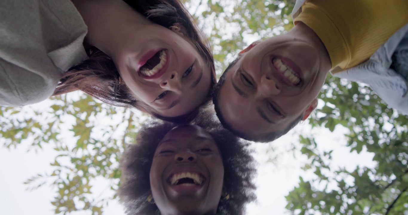 Diverse female friends leaning toward camera below, laughing and adjusting heads under leafy canopy