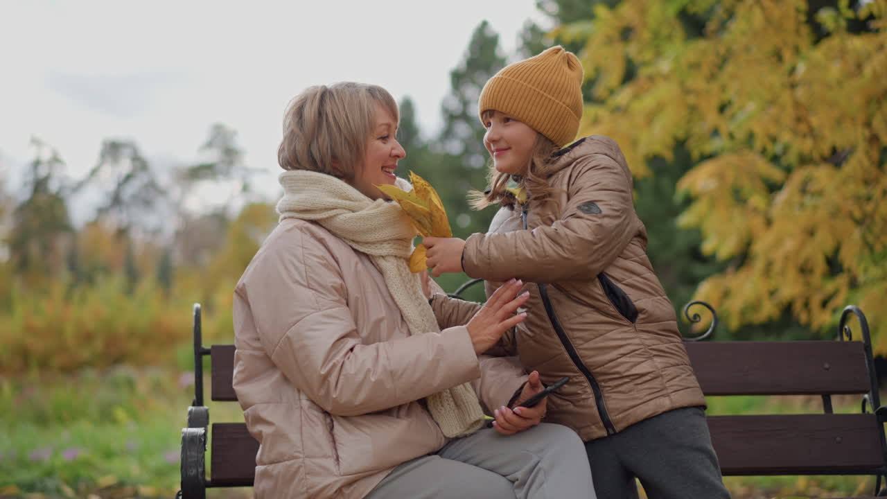 mother focused on phone as daughter approaches with bright yellow autumn leaf and warmly hugs in crisp park setting surrounded by golden falling foliage capturing joyful family moment