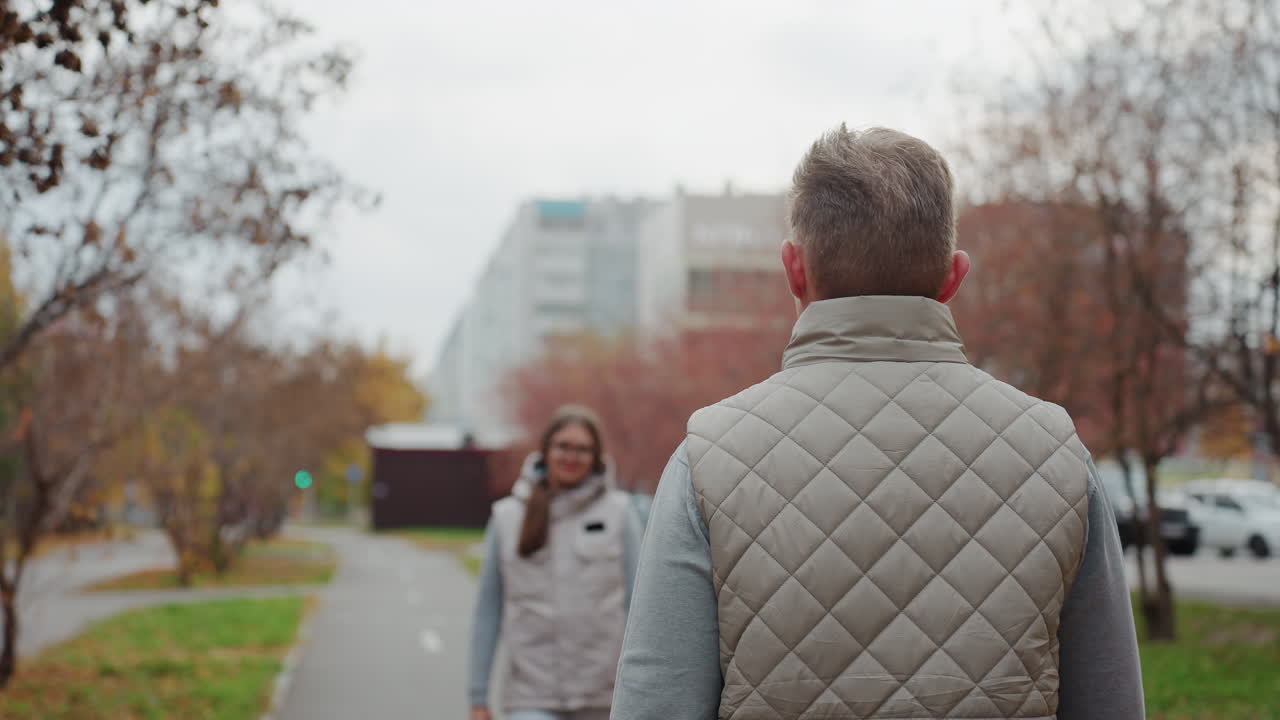 Young couple wearing same outfit walk toward each other and share warm hug in urban park as autumn leaves sway gently in wind with painted nails visible and modern office buildings in background