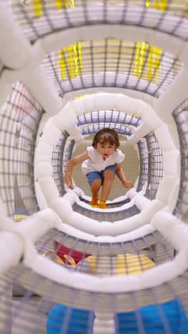 a happy and energetic little girl has fun climbing and crawling through a modern, futuristic-looking play tunnel at an indoor playground or kids' cafe - vertical slow motion