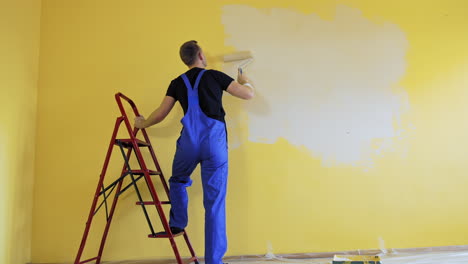 craftsman painting walls near the building ladder. professional painter in blue overalls doing a renovation in the room with a paint roller. rear view.