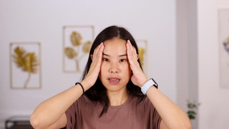 asian woman in casual clothing holds her head and face, expressing discomfort from a headache, in a brightly lit modern living room with framed art