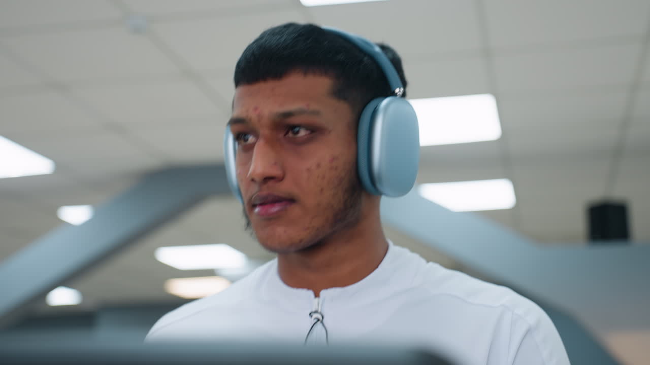 focused man wearing over-ear headphones and white athletic top works out in modern gym environment, looking sideways with concentration while ceiling lights cast bright glow over clean interior space