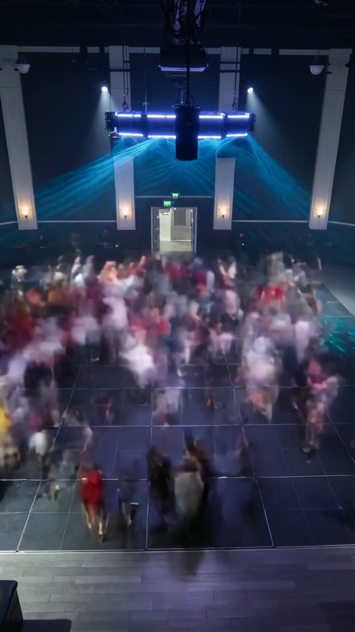 Moving crowd of partygoers wearing colorful clothes dancing on gridded dance floor, blue spotlights