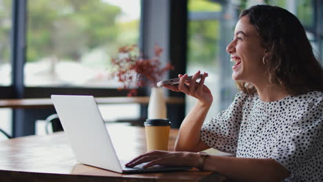 Joven-Empresaria-Sentada-Con-Una-Computadora-Portátil-En-Una-Cafetería-Tomando-Una-Llamada-Por-Teléfono-Móvil-Usando-Un-Micrófono