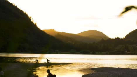 silhouette of people relaxing at riverside of mountain lake at sunset