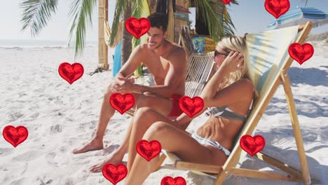 Animation-of-heart-digital-icons-over-couple-in-deckchairs-on-beach