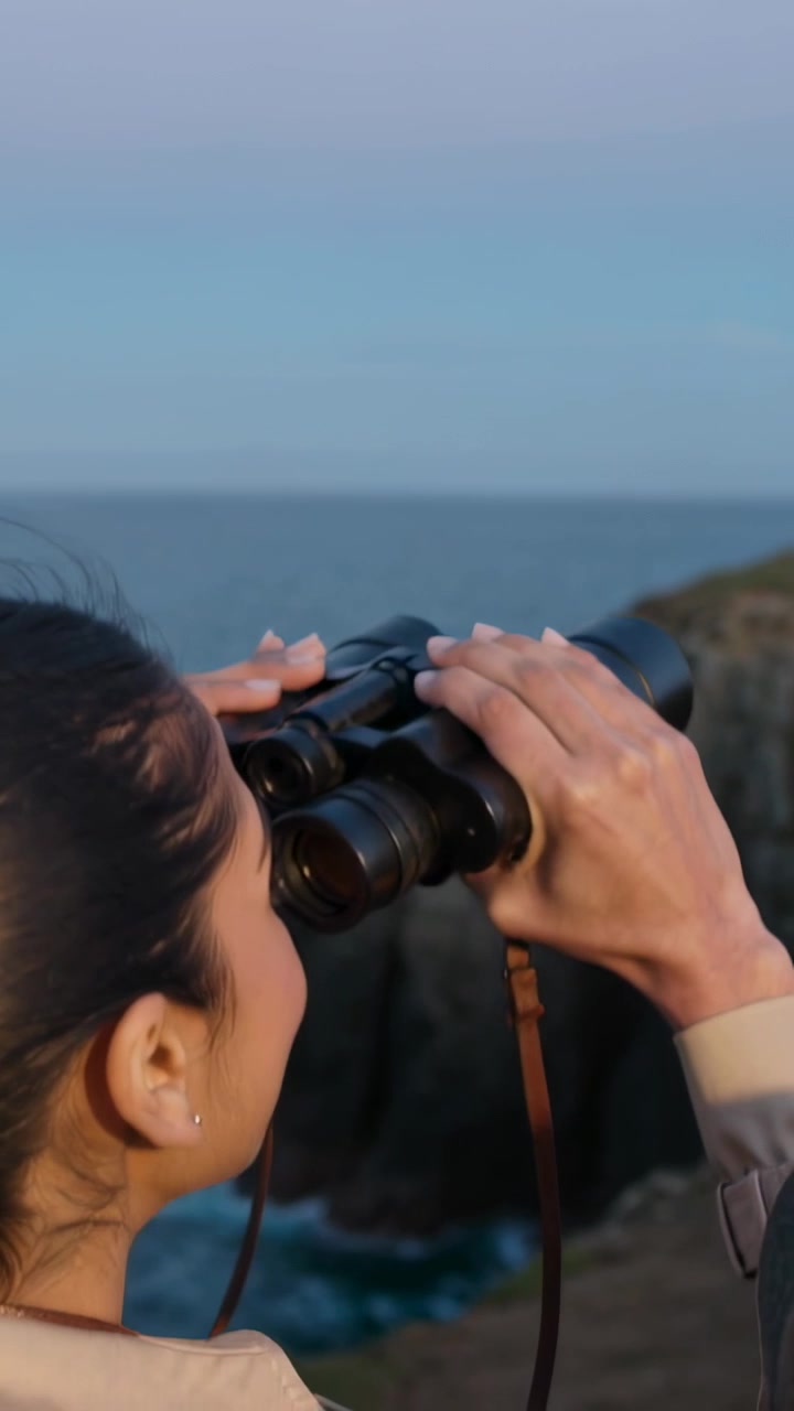 Vertical video: View opening woman in tan jacket raising binoculars to scan cliff, copy space
