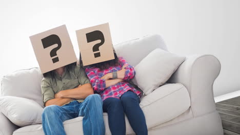 una pareja caucásica sentada en un sofá con cajas de cartón con signos de interrogación sobre sus cabezas