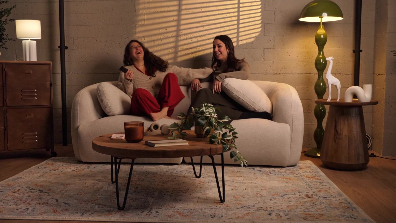 Two women sitting on a sofa watching TV, holding a remote control, pointing at screen and laughing together in a warm living room. Concept for streaming, friendship, and home entertainment.