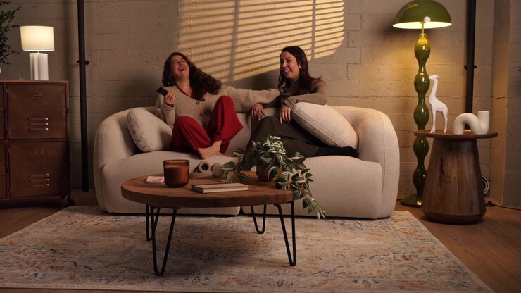 Two women sitting on a sofa watching TV, holding a remote control, pointing at screen and laughing together in a warm living room. Concept for streaming, friendship, and home entertainment.
