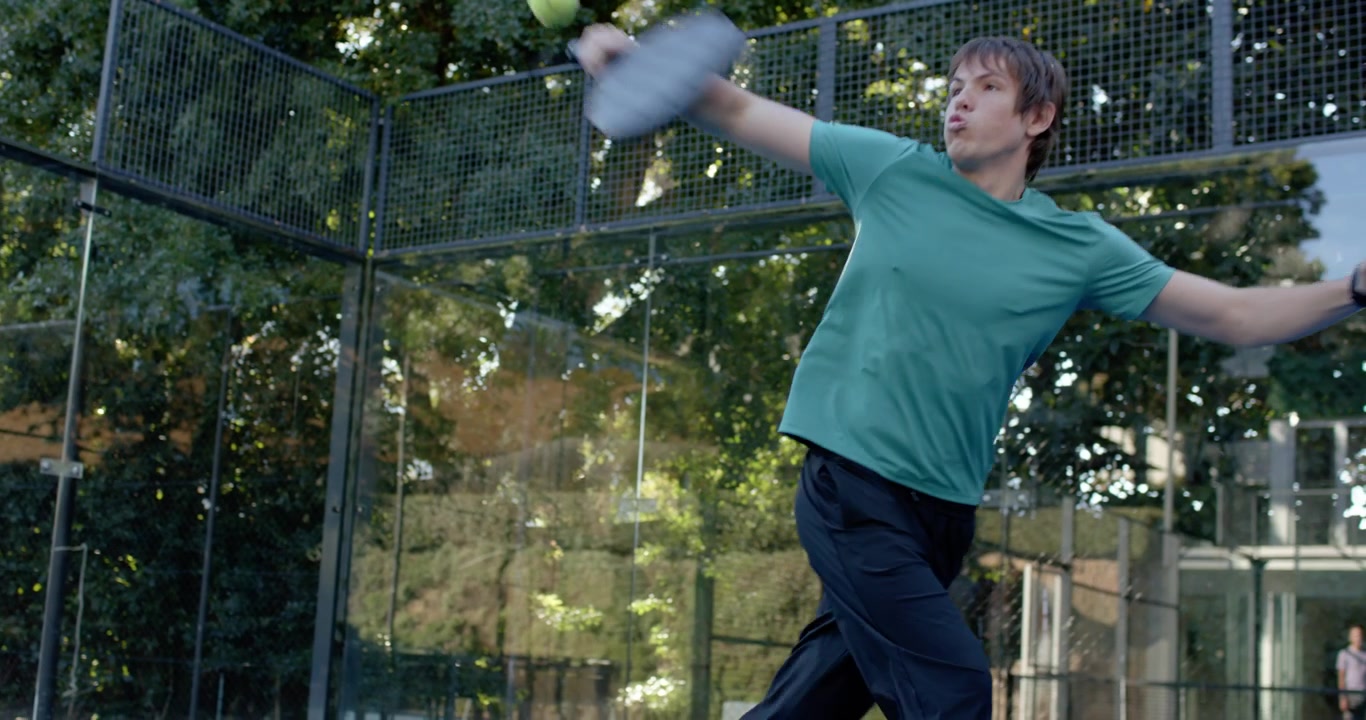 Adult male teal-shirted winding, swinging paddle, returning incoming lob at padel court, copy space