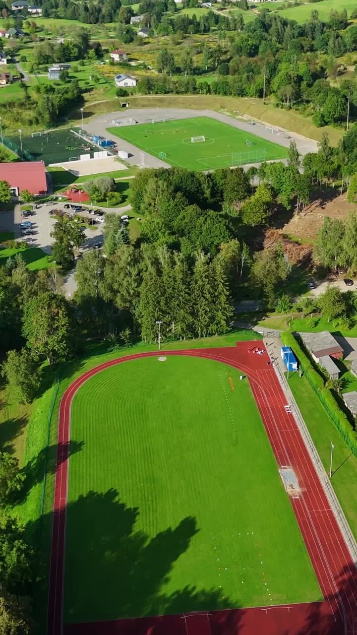 Vista aérea que revela la pista de atletismo, el campo de fútbol y los alrededores verdes de Talsi, Letonia, bajo cielos azules brillantes, destacando la infraestructura deportiva urbana, cámara de dron vertical inclinándose hacia abajo