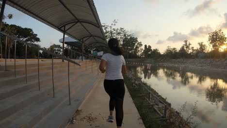 Una-Toma-En-Cámara-Lenta-De-Una-Joven-Tailandesa-Trotando-En-Una-Acera-Cerca-De-Un-Pequeño-Lago-Durante-La-Puesta-De-Sol,-Tailandia