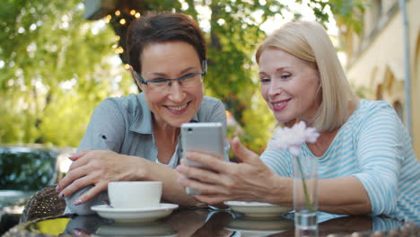 two women friends enjoying coffee and using a smartphone in an outdoor cafe