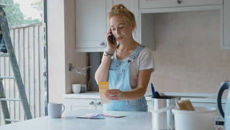 Woman-Renovating-Kitchen-At-Home-Looking-At-Paint-Swatches-And-Talking-On-Mobile-Phone