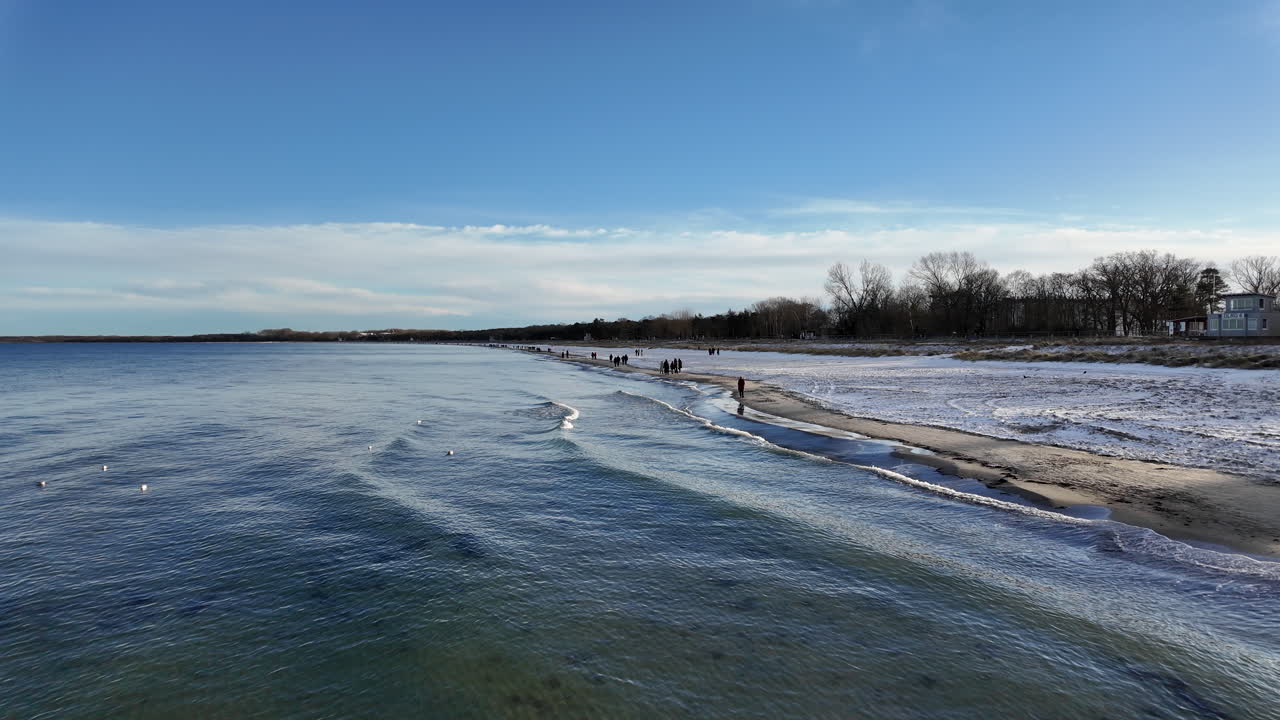 Winterliche Küstenlandschaft an der Ostsee in Boltenhagen, Deutschland, gefilmt im Januar. Das Video zeigt einen verschneiten Strand mit Menschen, die an der Küstenlinie unter einem dramatischen Winterhimmel entlanggehen.