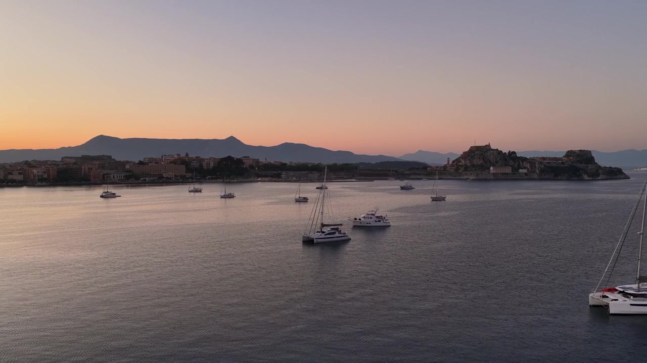 Veduta panoramica aerea del porto di Corfù al tramonto con barche a vela e catamarani ancorati che galleggiano sulle calme acque del Mar Mediterraneo, sullo sfondo della storica Vecchia Fortezza e montagne lontane.